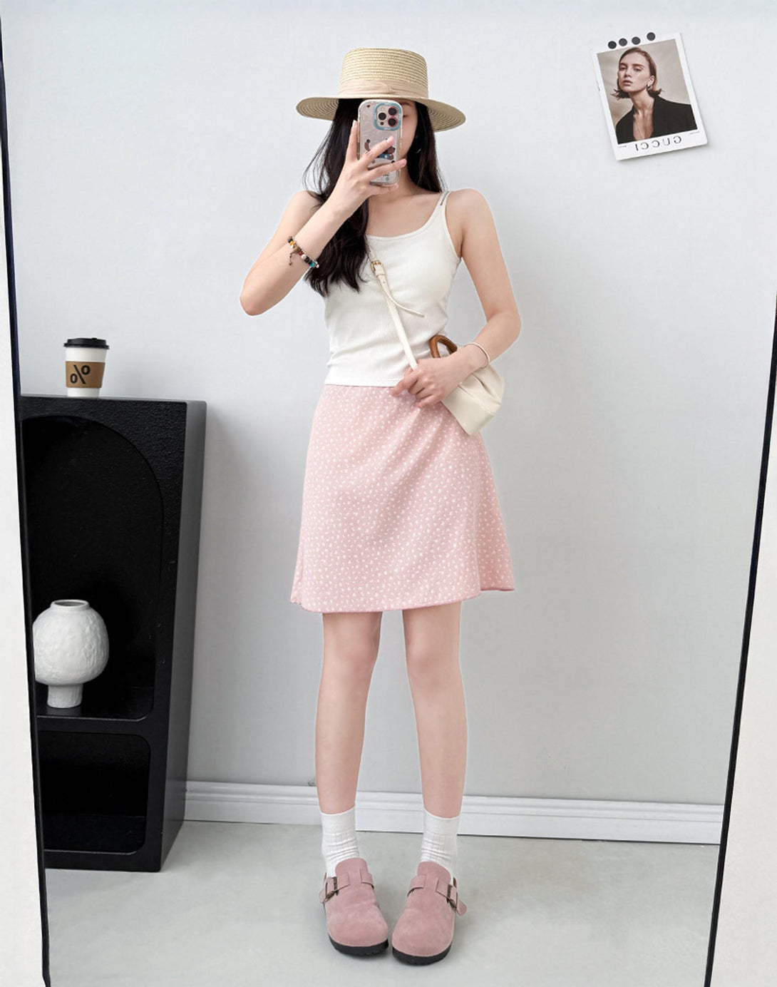 Woman taking a mirror selfie wearing a white top, bias cut feminine pink skirt, and pink shoes in a minimalistic room.